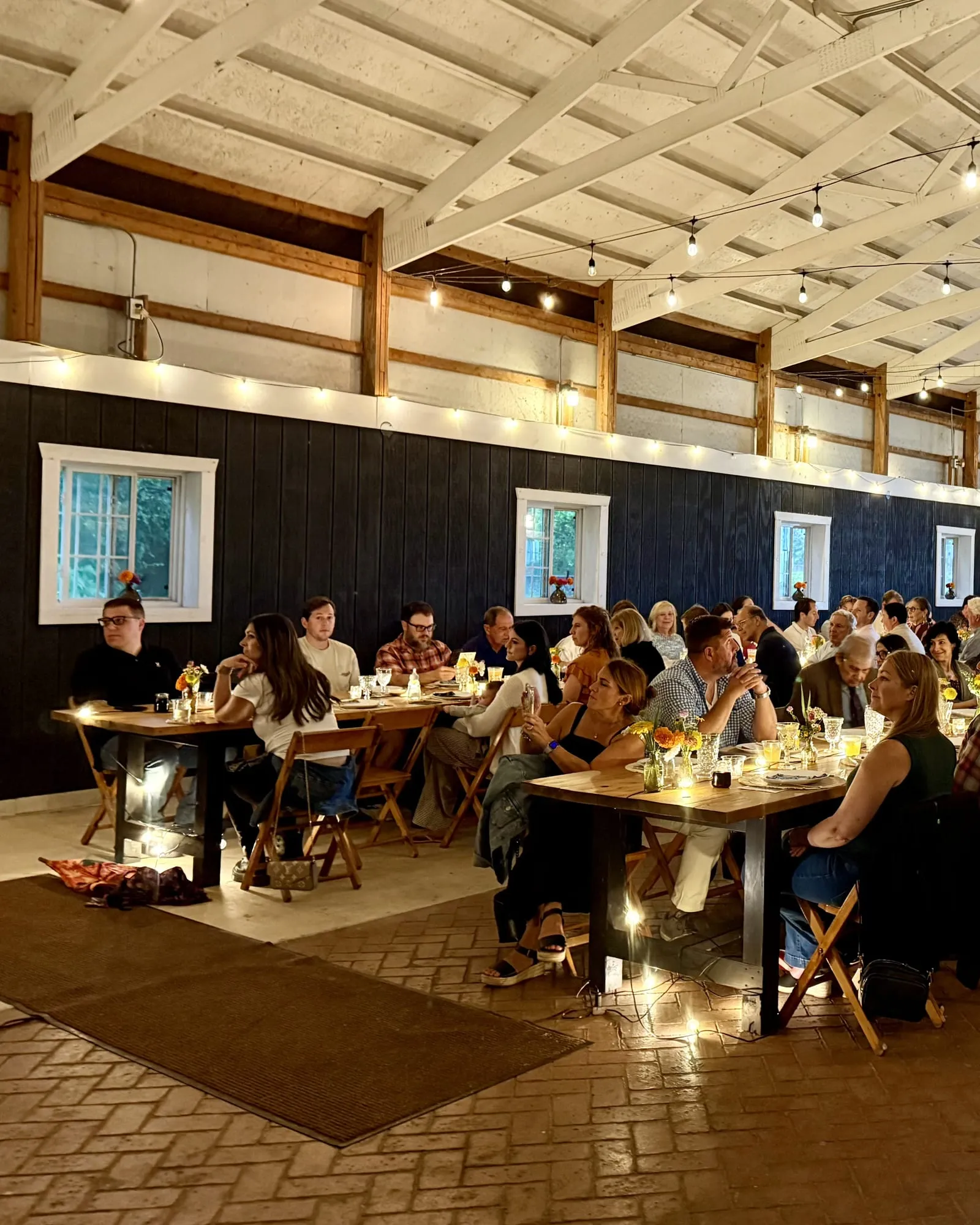 Guests seated at long farm tables under string lights in a barn