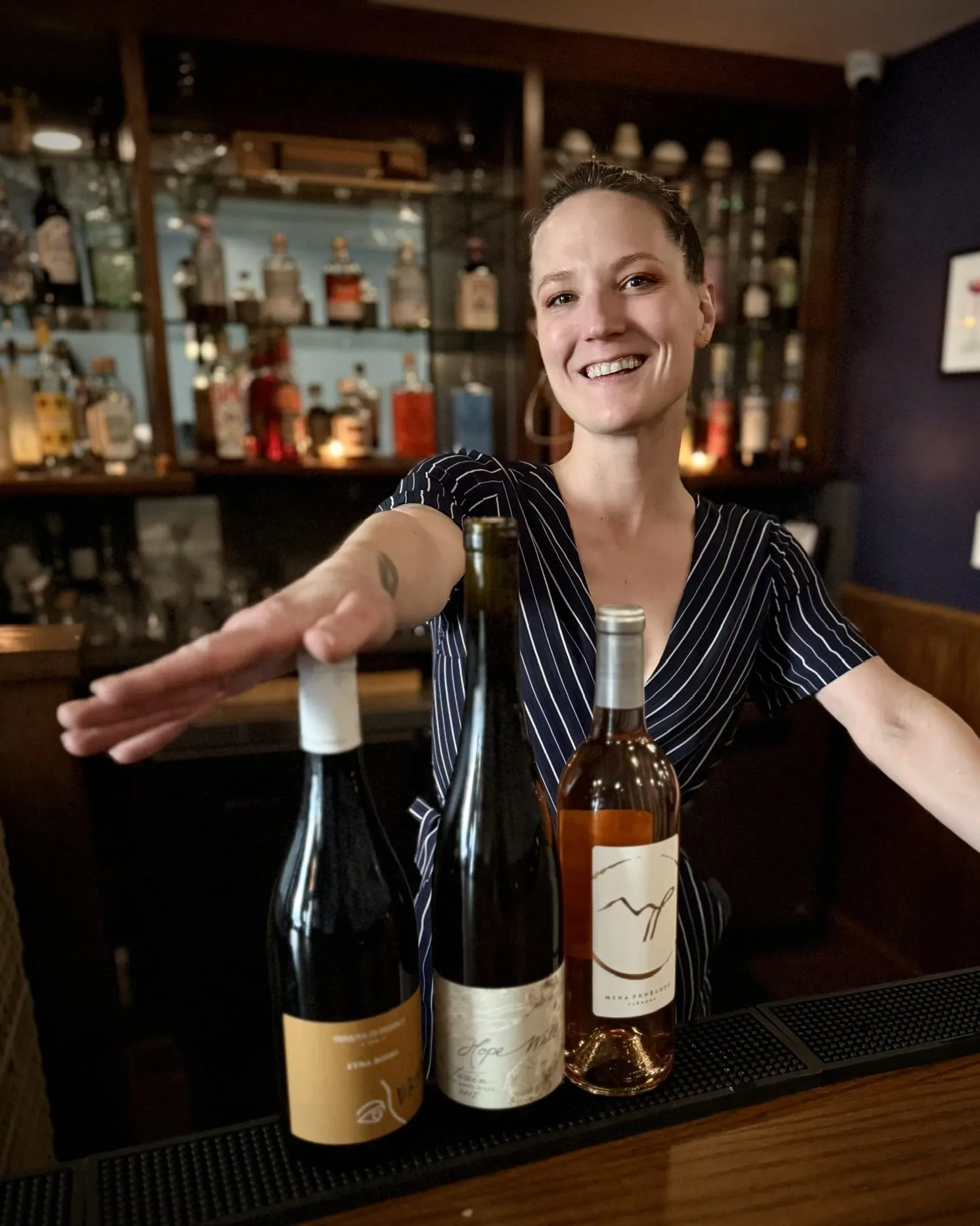 Ali smiling behind the bar, presenting three bottles for a tasting